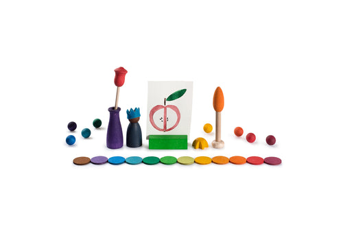 The image shows a collection of colorful wooden toys and objects arranged in a visually appealing manner. The objects include various shapes such as spheres, discs, and figures, all painted in vibrant colors. In the center, there is a small card with a drawing of a peach, held upright by a green stand. The arrangement of the objects and the use of bright colors make the image interesting and visually engaging.