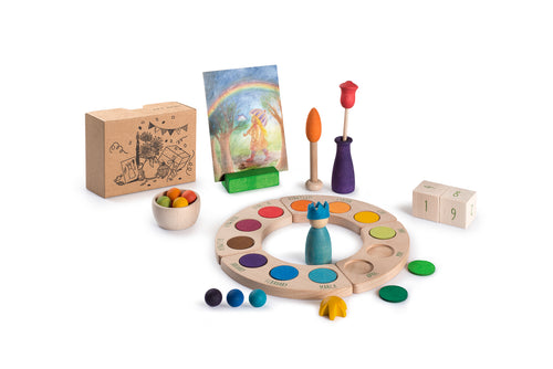 The image shows colorful wooden toys and objects arranged on a white surface. There is a circular wooden board divided into colored sections labeled with the months of the year. In the center, there is a small blue figurine. Surrounding the board, there are various wooden pieces including balls, a bowl with colored balls, a wooden flower in a vase, and dice with numbers. Additionally, there is a cardboard box with illustrations and a painting of a person under a rainbow.