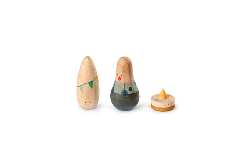 The image shows three wooden toys. On the left, there is an egg-shaped figure with a simple design, featuring a string of green and yellow triangular flags painted around its middle. In the center, there is another egg-shaped toy with a more detailed design, resembling a person with a painted face and clothing. On the right, there is a small spinning top with a conical tip and a circular base, decorated with small colored dots.