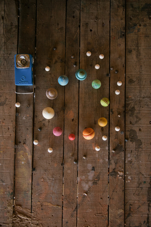 Colorful balls and white marbles arranged on a wooden surface, forming a spiral pattern. A blue Corona toy camera is placed on the left side of the arrangement.