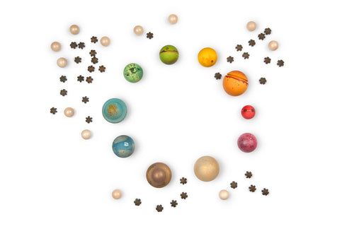 A circular arrangement of colorful wooden balls and star-shaped pieces on a white background. The balls are painted in various colors and patterns, resembling planets and celestial bodies, with some in blue, green, yellow, orange, red, and brown tones. Smaller beige wooden balls and dark brown star shapes surround the larger spheres, creating a look reminiscent of a solar system with orbiting elements